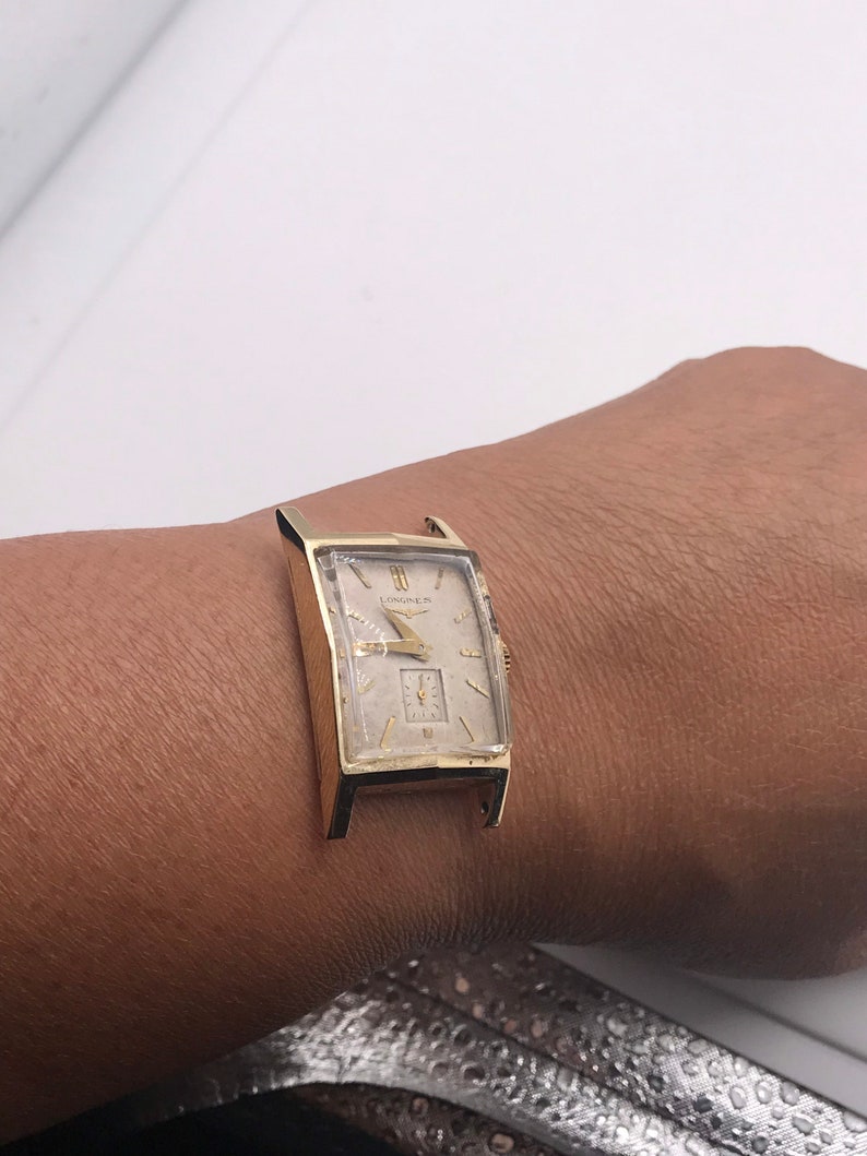 May include: A gold-toned rectangular wristwatch with a cream-colored face. The watch features a small subdial and the brand name "Longines" is visible. The watch is on a person's wrist.