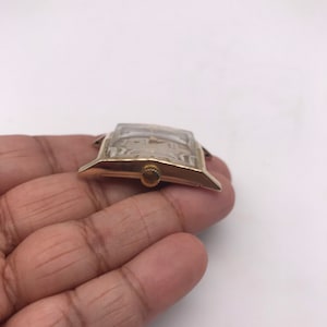 May include: A gold-toned rectangular wristwatch with a light-colored face. The watch has a small, round crown and a clear crystal. The watch is held in a hand against a white background.