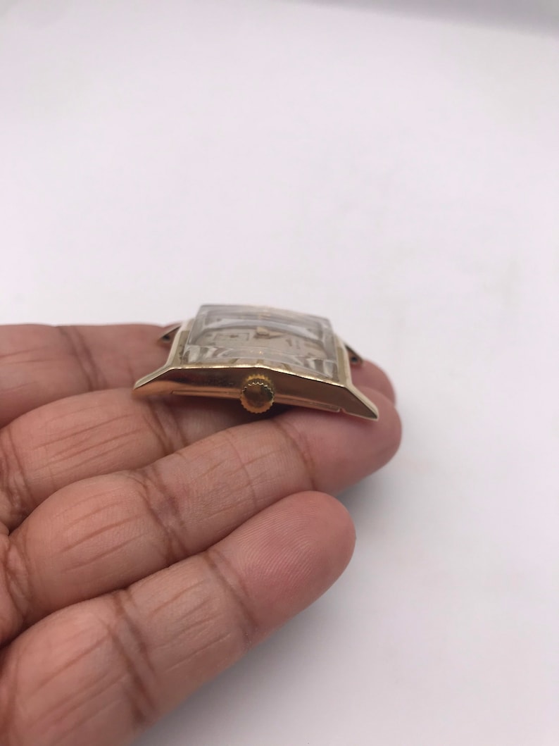 May include: A gold-toned rectangular wristwatch with a light-colored face. The watch has a small, round crown and a clear crystal. The watch is held in a hand against a white background.
