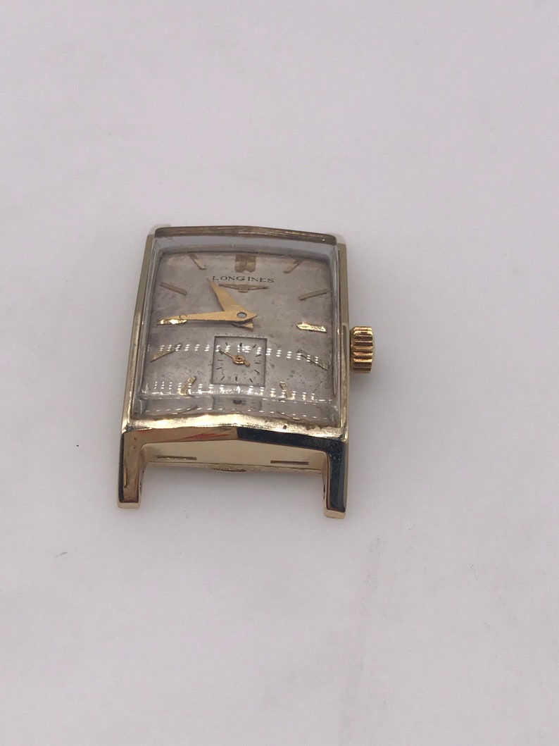 May include: A vintage rectangular Longines watch with a gold-tone case and a silver-tone face. The watch features gold hands and hour markers, and a small subdial. The Longines logo is visible at the top of the watch face.