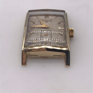 May include: A vintage gold-toned Longines wristwatch with a rectangular face. The watch features a silver-colored dial with gold hands and hour markers. The watch has a small subdial and a gold crown. The watch is displayed on a white surface.