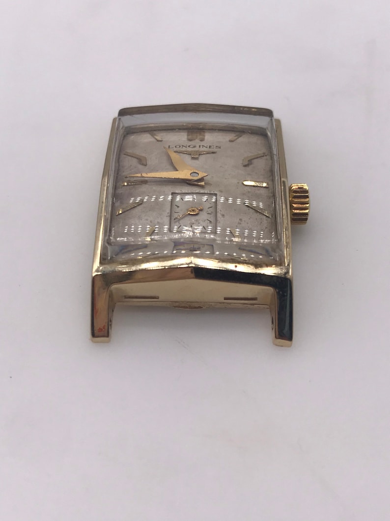 May include: A vintage gold-toned Longines wristwatch with a rectangular face. The watch features a silver-colored dial with gold hands and hour markers. The watch has a small subdial and a gold crown. The watch is displayed on a white surface.