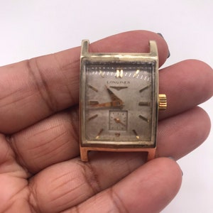 May include: A vintage gold-toned Longines watch with a rectangular face. The watch features a small seconds dial and gold hands. The watch is held in a hand against a white background. The watch face has a light-colored background with the Longines logo.