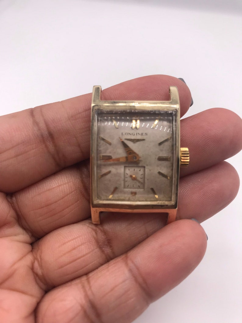 May include: A vintage gold-toned Longines watch with a rectangular face. The watch features a small seconds dial and gold hands. The watch is held in a hand against a white background. The watch face has a light-colored background with the Longines logo.