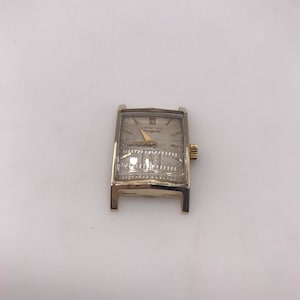 May include: A vintage gold-toned rectangular wristwatch with a light-colored face. The watch features a gold-colored band and a small, gold-colored crown. The watch face has the brand name "LONGINES" printed on it.
