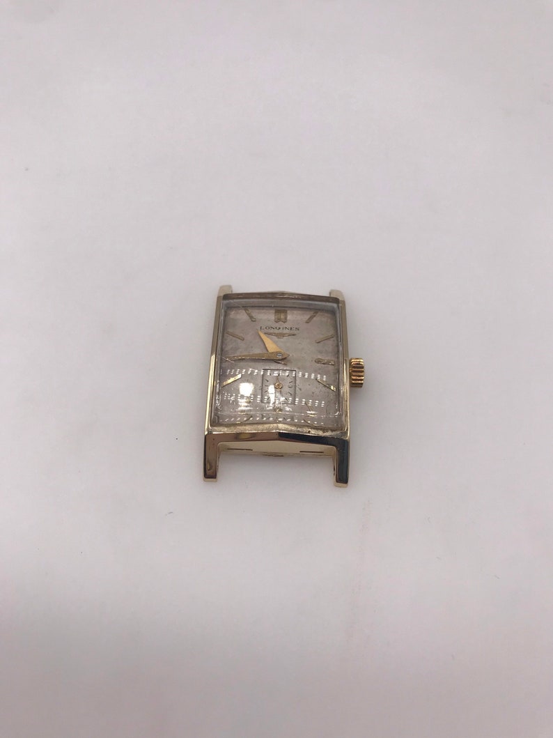 May include: A vintage gold-toned rectangular wristwatch with a light-colored face. The watch features a gold-colored band and a small, gold-colored crown. The watch face has the brand name "LONGINES" printed on it.