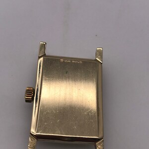 May include: A gold-toned rectangular watch case with a textured surface. The watch has a crown on the side and lugs for attaching a band. The text "14K GOLD" is visible on the watch.