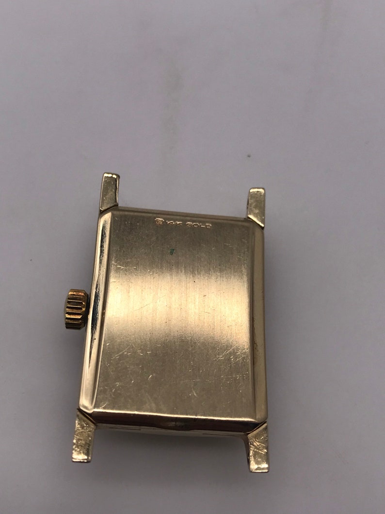 May include: A gold-toned rectangular watch case with a textured surface. The watch has a crown on the side and lugs for attaching a band. The text "14K GOLD" is visible on the watch.