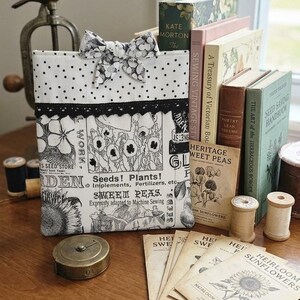 May include: A rectangular fabric pouch featuring a black and white botanical print with text reading Seeds! Plants! and Sweet Peas, accented by a black lace trim and a polka dot bow. The pouch is displayed alongside vintage books and seed packets.