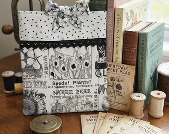 May include: A rectangular fabric pouch featuring a black and white botanical print with text reading Seeds! Plants! and Sweet Peas, accented by a black lace trim and a polka dot bow. The pouch is displayed alongside vintage books and seed packets.