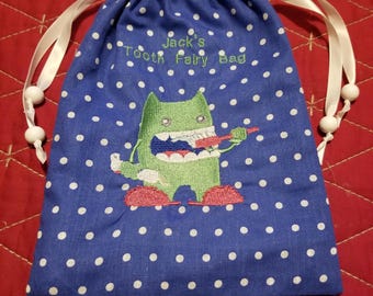 May include: Blue tooth fairy bag with white polka dots and an embroidered green monster with red shoes. The bag has white drawstrings and white beads. The bag is personalized with the text "Jack's Tooth Fairy Bag".