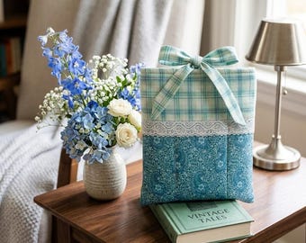 May include: A blue floral book sleeve with a plaid top and a white lace trim, topped with a matching bow. It sits on a vintage book, beside a vase of blue and white flowers, on a wooden table.