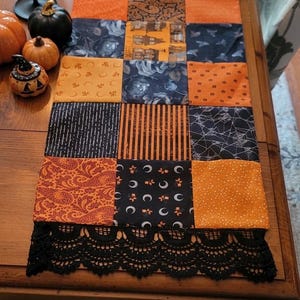 May include: A rectangular patchwork table runner featuring a variety of Halloween-themed fabric squares in orange, black, and white. The runner is finished with a decorative black lace trim on both ends, showcasing patterns of pumpkins, moons, and webs.