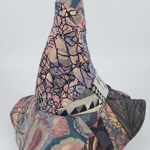 May include: A pointed hat constructed from various panels of woven fabric featuring a mix of textures and patterns. The design includes shades of pink, blue, and grey with a prominent band around the base displaying a black and white geometric triangle motif.