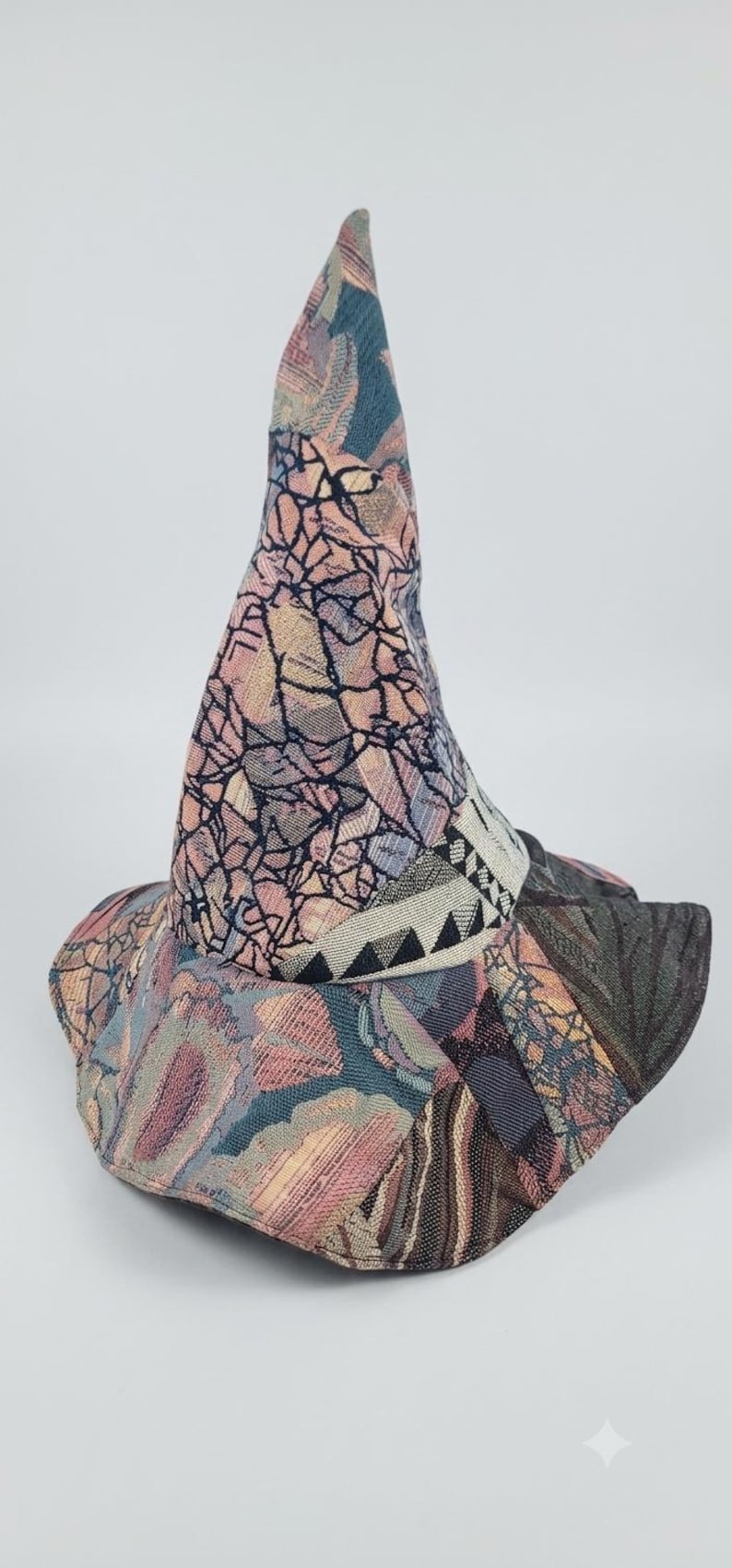 May include: A pointed hat constructed from various panels of woven fabric featuring a mix of textures and patterns. The design includes shades of pink, blue, and grey with a prominent band around the base displaying a black and white geometric triangle motif.