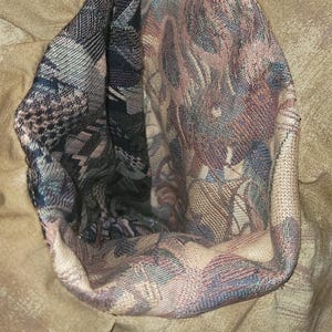 May include: A tan fabric garment featuring a deep, open pocket or interior section lined with a multi-colored woven tapestry fabric. The lining displays a complex pattern of dark blue, muted red, and beige threads in a dense, textured weave.