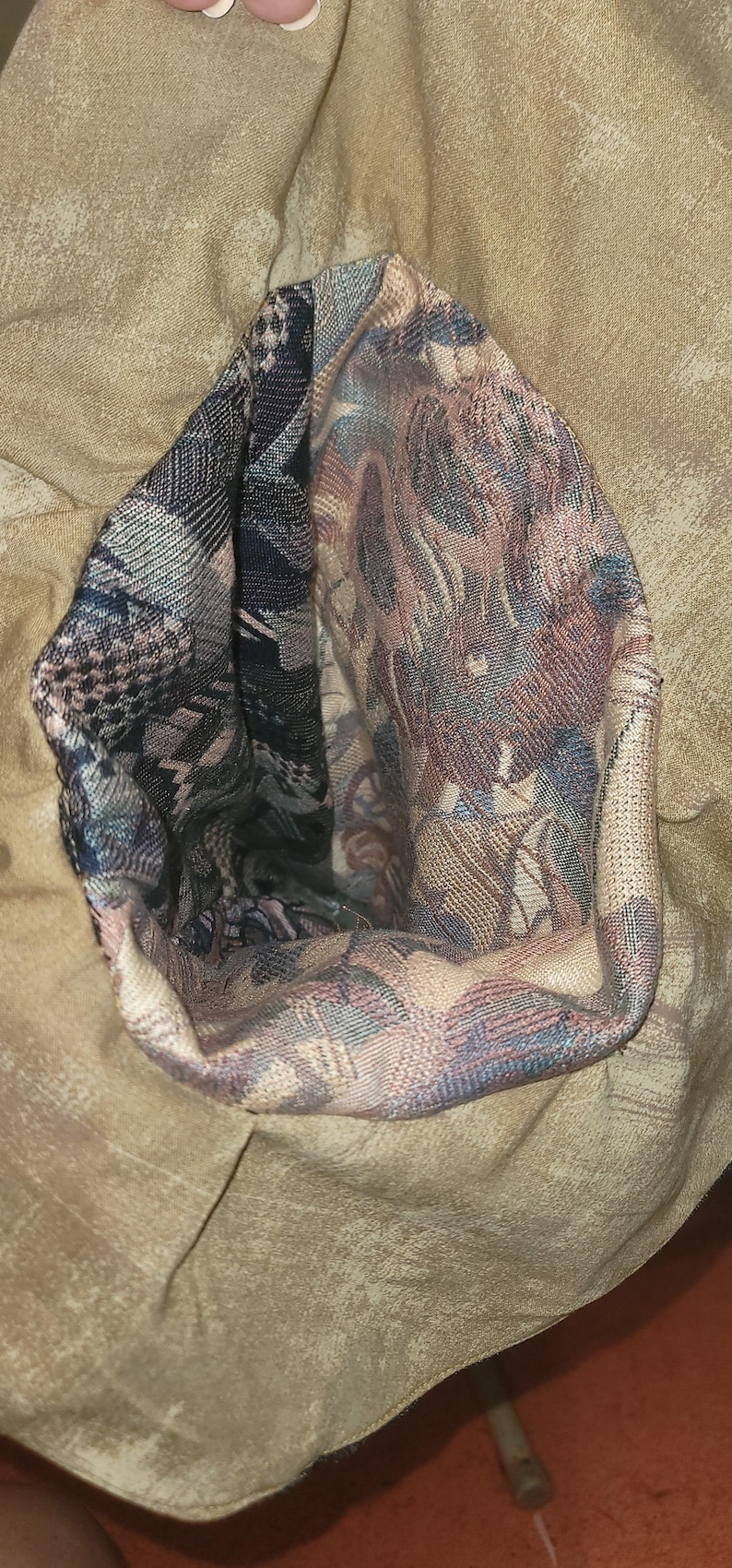 May include: A tan fabric garment featuring a deep, open pocket or interior section lined with a multi-colored woven tapestry fabric. The lining displays a complex pattern of dark blue, muted red, and beige threads in a dense, textured weave.