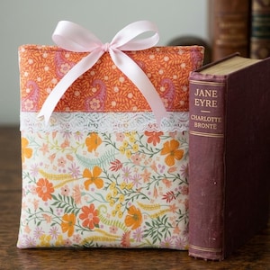 May include: A floral fabric book sleeve with a pink ribbon bow. The sleeve features a paisley print on top and a floral pattern on the bottom, separated by a white lace trim. A maroon book with the title "Jane Eyre" is next to the sleeve.