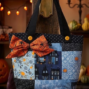 May include: A patchwork tote bag featuring a variety of blue and black fabric squares with patterns of houses, spiderwebs, and pumpkins. The bag has black handles, two yellow buttons, and a large orange bow with a black spiderweb print attached.