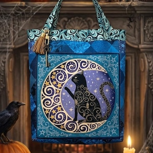May include: A blue fabric tote bag featuring a central circular graphic of a black cat sitting inside a crescent moon surrounded by stars. The bag has a patterned top border, two handles, and a small gold-toned key charm with a brown tassel attached.