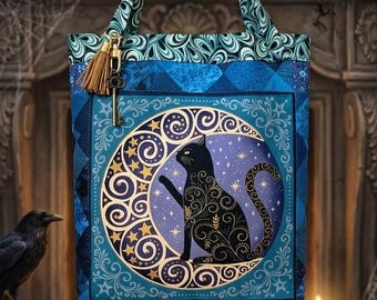 May include: A blue fabric tote bag featuring a central circular graphic of a black cat sitting inside a crescent moon surrounded by stars. The bag has a patterned top border, two handles, and a small gold-toned key charm with a brown tassel attached.