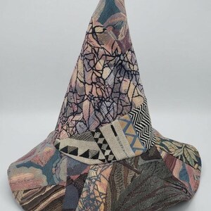 May include: A pointed hat constructed from various fabric patches featuring a mix of textures and patterns. The design includes muted tones of blue, pink, and grey, with a band around the base showcasing geometric black and white herringbone and triangle motifs.
