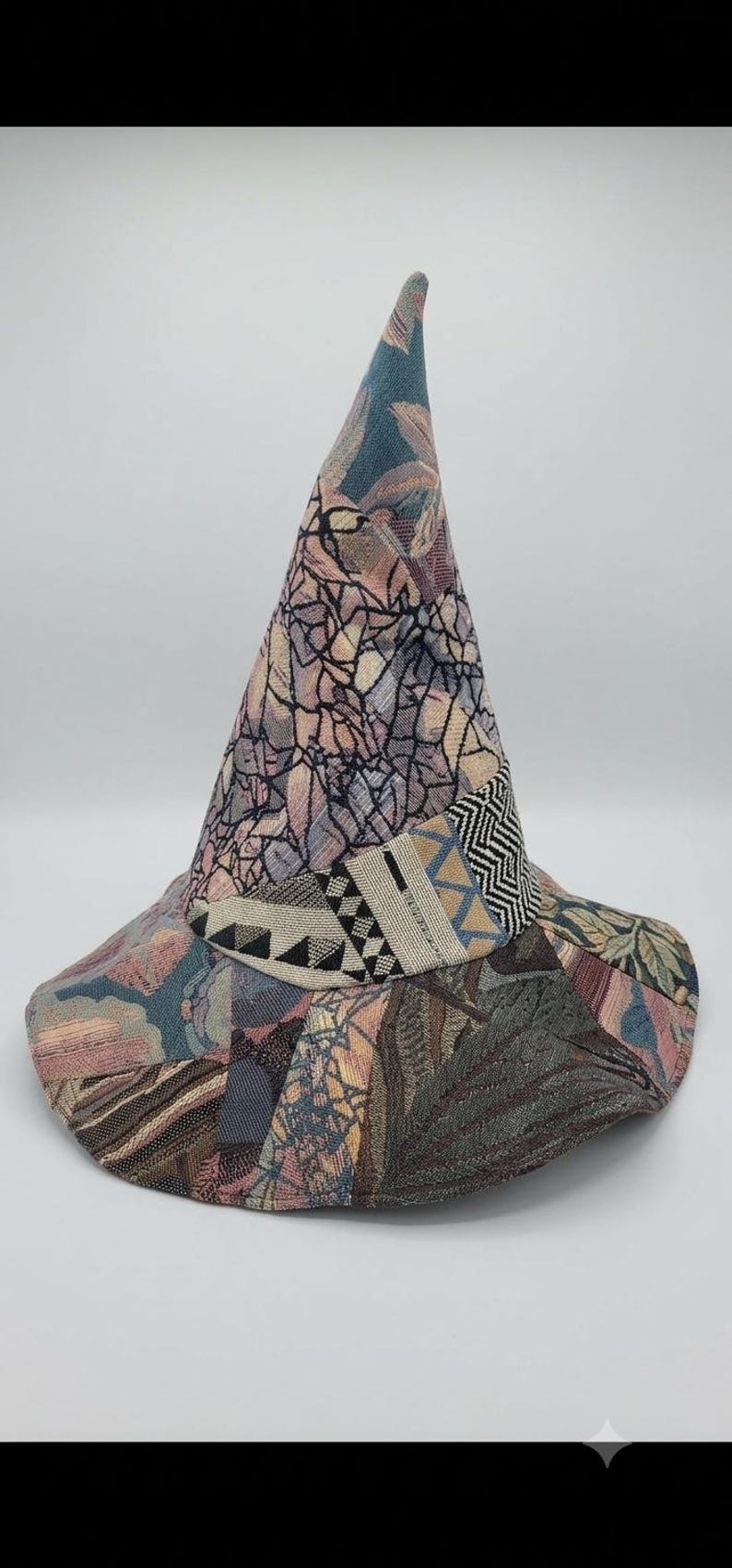 May include: A pointed hat constructed from various fabric patches featuring a mix of textures and patterns. The design includes muted tones of blue, pink, and grey, with a band around the base showcasing geometric black and white herringbone and triangle motifs.