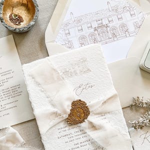 May include: Elegant wedding invitation suite featuring a cream-colored invitation with a gold wax seal and ribbon. Includes a matching envelope with a detailed illustration, a ring in a box, and a details card. The suite is perfect for a formal wedding.
