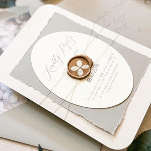 May include: A wedding invitation suite featuring a cream-colored card with an oval insert and a gray paper backing. The insert reads "Kindly Reply" and includes a QR code. A wax seal with a floral design secures the card, tied with twine.