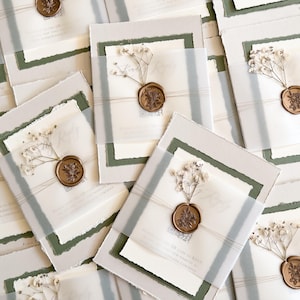 May include: An assortment of wedding invitations with a layered design. Each invitation features a white card with torn edges, a translucent overlay, and a sprig of dried flowers, secured with a bronze wax seal.