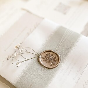 May include: Elegant wedding invitation suite featuring a gold wax seal with a floral design. The invitation is tied with a white ribbon and adorned with delicate white flowers. The paper has a textured, off-white appearance.