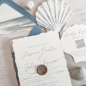 May include: Elegant wedding invitation suite with a beach theme. Includes invitation, RSVP card, and envelope with a watercolor beach scene. The invitation is tied with twine and sealed with a seashell wax seal. Seashell accents complete the design.