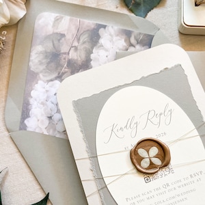 May include: Elegant wedding invitation suite featuring a gray and white color scheme. The invitation includes a floral-lined envelope, a reply card with a wax seal, and delicate twine. The text "Kindly Reply" is visible.
