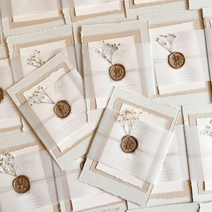 May include: Wedding invitation suite featuring layered paper in cream and beige tones. Each invitation is sealed with a gold wax stamp and adorned with delicate white flowers. The suite includes a reply card and vellum wrap.