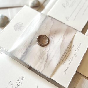 May include: Elegant wedding invitations with a beach theme. The invitations feature a watercolor design, a wax seal, and the text "Dinner and dancing to follow". The invitations are on textured paper.