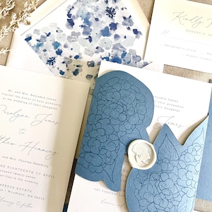 May include: Elegant wedding invitation suite featuring a light blue floral design. Includes invitation cards, RSVP cards, and a blue die-cut card with a wax seal. The invitation text includes names, dates, and location details.