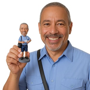 May include: A bobblehead figurine of a postal worker, in a blue uniform and holding mail, is held up by a smiling man in a blue shirt. The figurine has a black satchel and stands on a grey base. The man has grey hair and a beard.