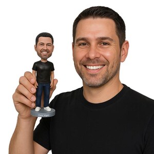 May include: A bobblehead figurine of a man wearing a black t-shirt and blue jeans, standing on a gray base. The base has the name "Conr Vigen" printed on it. The figurine is held by a person wearing a black t-shirt.
