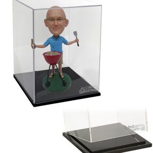 May include: A custom bobblehead of a man in a blue shirt and khaki shorts, holding a spatula and a hot dog. The figure stands in front of a red grill on a green base, inside a clear display case. The base is black.
