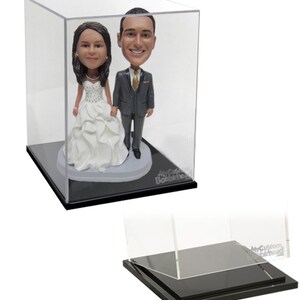 May include: A clear acrylic display case with a black base, showcasing two custom bobbleheads of a bride and groom. The bobbleheads are standing on a small platform inside the case.