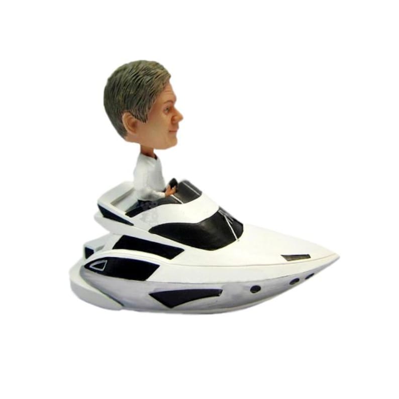May include: A white and black yacht figurine with a bobblehead of a person with grey hair. The figurine has a detailed design, with a person in a white shirt at the helm. The yacht has black accents and a sleek design.