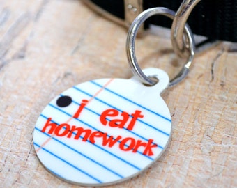 May include: A white round dog tag with blue and red lines, a black dot, and the text "i eat homework".