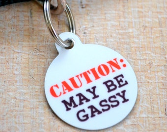 May include: White round dog tag with a black strap. The tag has the text "CAUTION: MAY BE GASSY" printed in red and black.