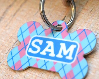 May include: A blue and pink bone-shaped dog tag with a silver ring. The tag has a blue and white rectangle with the name "SAM" printed in white.