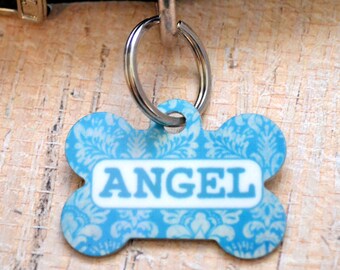 May include: A blue bone-shaped dog tag with a white damask pattern and the word "ANGEL" printed in white letters.