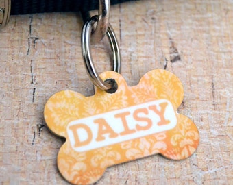 May include: A yellow bone-shaped dog tag with a white floral pattern and the name "DAISY" printed in white letters.