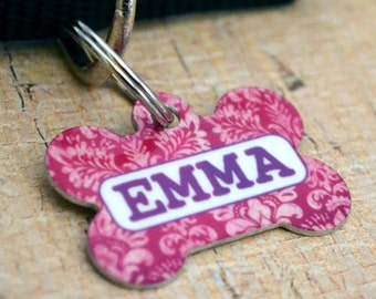 May include: Pink bone-shaped dog tag with a white and purple damask pattern and the name "EMMA" printed in purple letters.