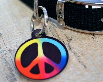 May include: A colorful peace sign dog tag with a rainbow design. The tag is attached to a black nylon dog collar with a silver buckle.