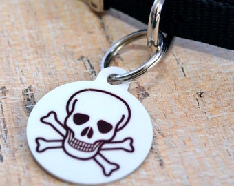 May include: White round dog tag with a black and white skull and crossbones design. The tag is attached to a black dog collar.