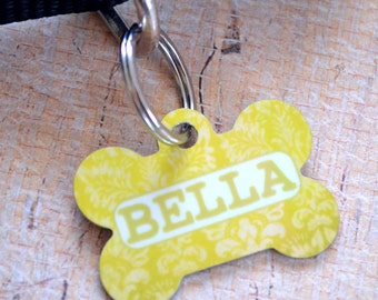 May include: Yellow bone-shaped dog tag with a white damask pattern and the name "BELLA" printed in white.